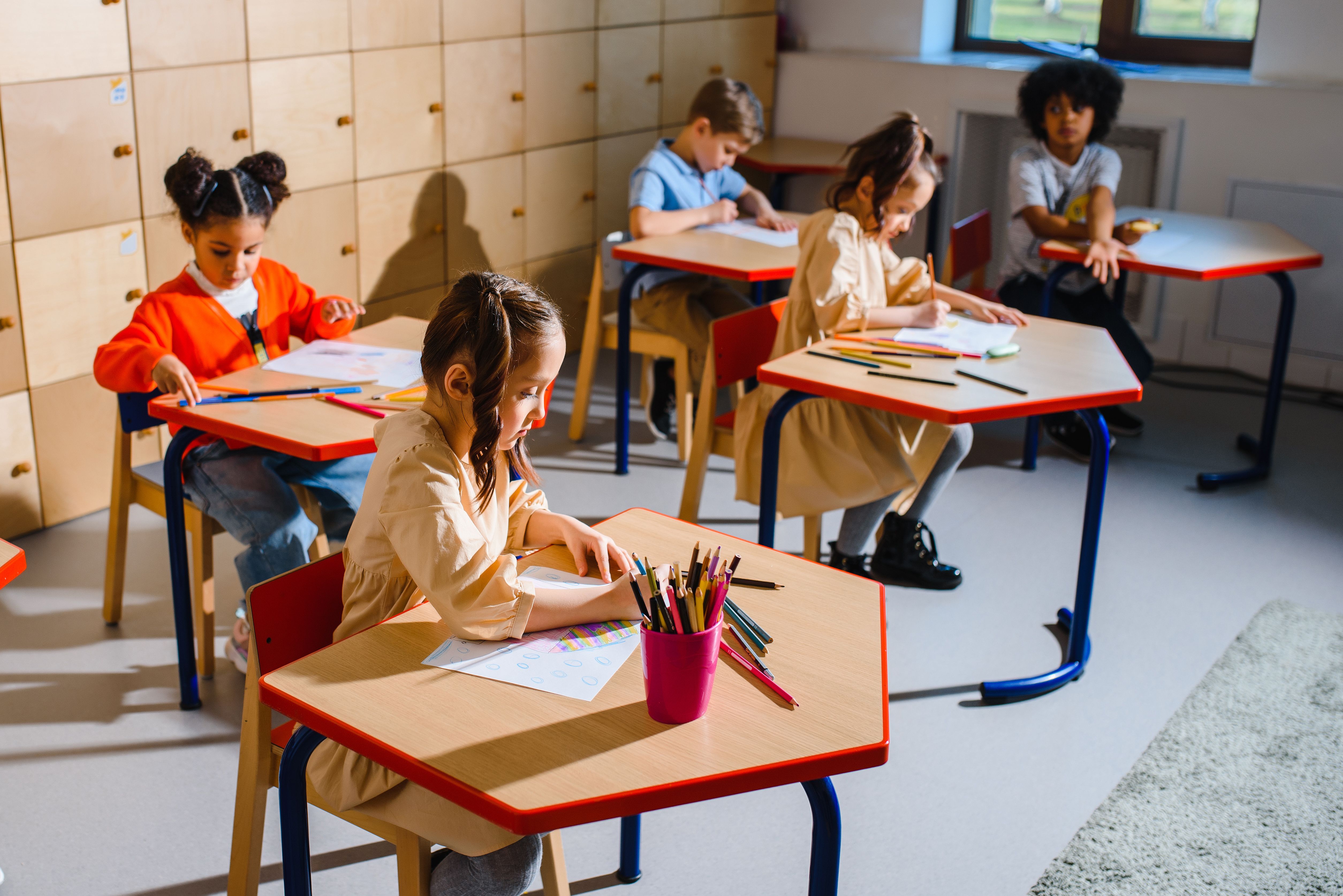 A group of children seated at desks in a bright classroom drawing and working with colored pencils